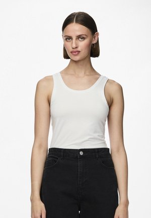 Kvinna med rakt brunt hår står och bär en vit ärmlös topp och svart byxa med hög midja mot en enkel vit bakgrund.