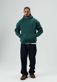 Bosgroene hoodie met een zak aan de voorkant, een losse pasvorm en geribbelde manchetten, gecombineerd met donkerblauwe jeans en bruine veterlaarzen.