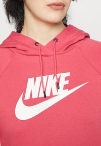Růžová mikina Nike s kapucí a šňůrkami, s velkým bílým logem. Měkká látka, raglánové rukávy a žebrované lemy na rukávech a spodním okraji.