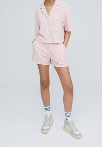 Camicia rosa a maniche corte con bottoni e tasca, abbinata a pantaloni corti coordinati. Persona indossa sneaker bianche con accenti beige e grigi, calzini a righe.