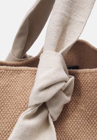 Sac fourre-tout en toile avec un corps marron texturé et des anses nouées beige clair. Présente un motif tissé et un matériau doux et durable.