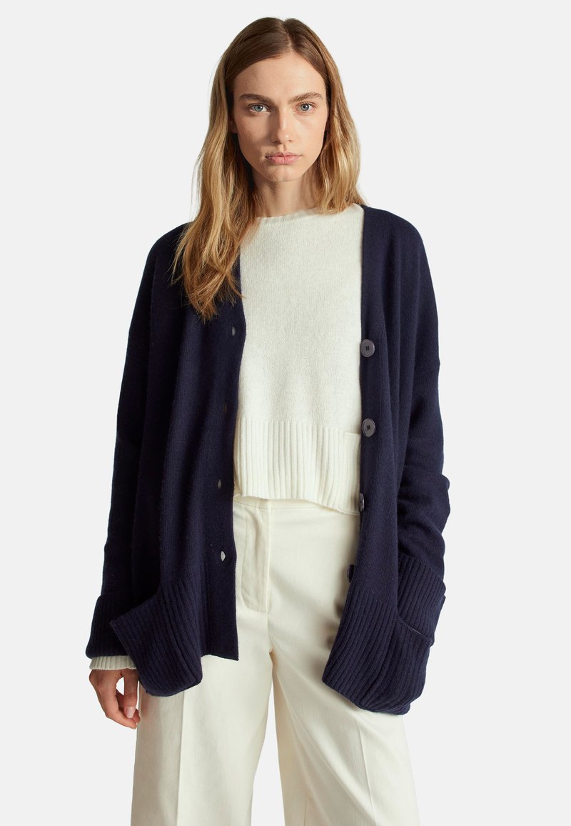 Námořnický cardigan s ribovanými manžetami a detaily kapes, se pěti knoflíky. Nosený přes svetřík v světle krémové barvě a v kombinaci s krémovými kalhotami.