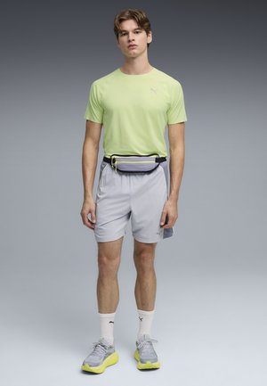 Ung mand står iført en lysegrøn kortærmet skjorte, grå shorts, grå og gule sneakers, hvide sokker og en grå bæltetaske med gul lynlås.