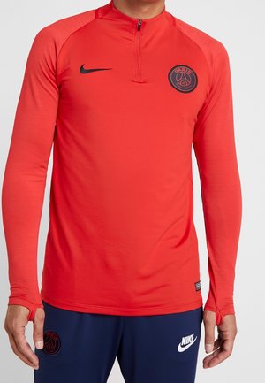 Mężczyzna ubrany w czerwoną długorękawową bluzę treningową Paris Saint-Germain z otworami na kciuki oraz granatowe sportowe spodnie Nike.