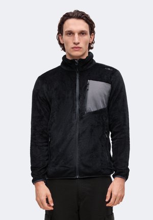 Un homme pose avec une veste polaire noire à col montant, dotée d’une fermeture éclair sur le devant et d’une poche poitrine grise zippée, sur un fond clair et uni.