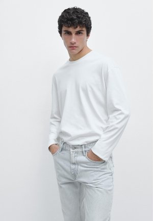 Jeune homme aux cheveux bouclés noirs portant une chemise blanche unie à manches longues et un jean gris clair, les mains dans les poches, debout devant un fond blanc.