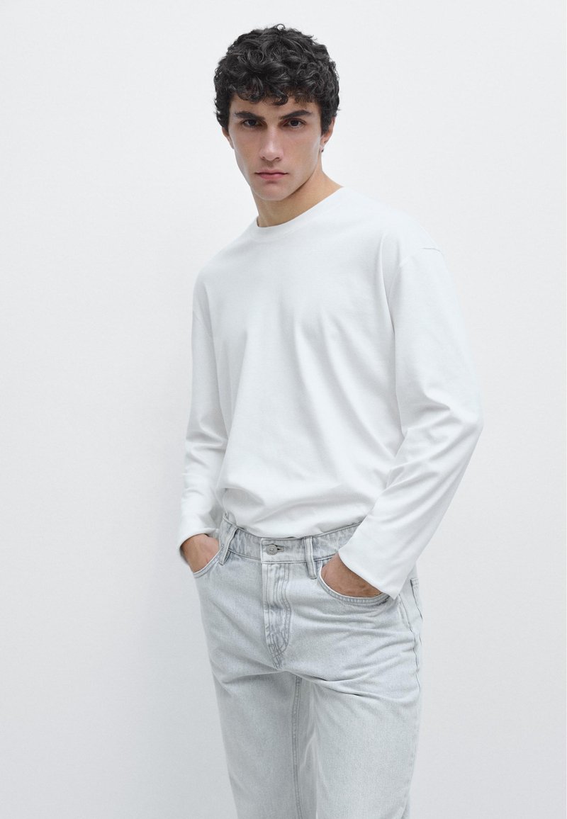 Junger Mann mit dunklem, lockigem Haar, trägt ein schlichtes weißes Langarmshirt und hellgraue Jeans, Hände in den Taschen, steht vor einem weißen Hintergrund.