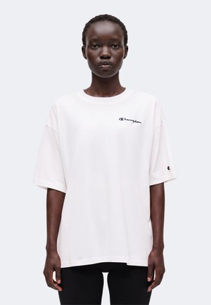 Person står vendt fremad iført en hvid oversize Champion T-shirt med logoer på bryst og ærme, kombineret med sorte bukser.