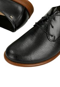Scarpe in pelle nera con design perforato, punta rotonda e suole in gomma marrone. Una scarpa ha una parte superiore liscia; l'altra presenta un sistema di allacciatura.
