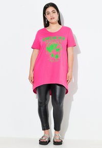 Roze t-shirt met groene "DISCO NIGHTCLUB" grafiek, ronde hals, korte mouwen, langere achterkant, gecombineerd met zwarte leggings en zilveren sandalen.