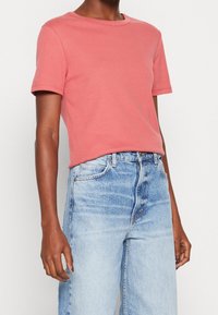 Person som bär en kortärmad korallrosa t-shirt instoppad i ljusblå högmidjade jeans mot en helt vit bakgrund.