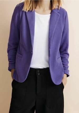 Donna che indossa un blazer viola sopra una camicia bianca e pantaloni neri a vita alta con le mani nelle tasche, su sfondo neutro.
