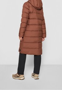 Brun isolerad pufferjacka med huva, med horisontella sömmar och en något längre nederkant. Bärs över svarta byxor och beiga skor.