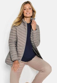 Glimlachende vrouw in een beige gewatteerde jas over een marineblauwe hoodie en beige broek, zittend tegen een effen witte achtergrond.