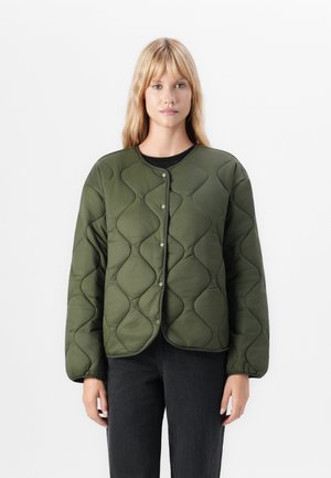 Frau mit schulterlangen blonden Haaren, die eine olivgrüne, gesteppte, knopfgeschlossene Jacke und schwarze Hosen trägt, steht vor einem weißen Hintergrund.