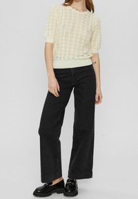 Pletený svetřík světle žluté barvy s texturovaným vzorem listů a vlnitým výstřihem, kombinovaný s černými širokými džíny s vysokým pasem a černými botami.