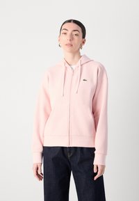 Hoddie rose clair à fermeture éclair avec une poche avant, des cordons et un petit logo vert. Présente des poignets côtelés et une texture lisse. À associer avec un jean noir.