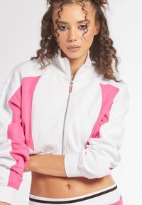 Giacca bianca accorciata con pannelli laterali rosa e chiusura con zip. Presenta un colletto alto e polsini a coste. Tessuto morbido con superficie liscia.