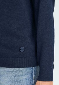 Mørkeblå striktrøje lavet af letvægtsmateriale, med en tekstureret overflade og en lille logo-knap nær kanten.