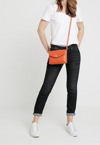 Orange crossbody bag with a flap closure and metal hardware, made of smooth leather. The model wears a white t-shirt and rolled black jeans.