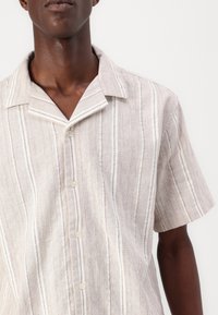 Camisa de botones de manga corta en beige claro con rayas verticales blancas. Presenta un cuello tipo solapa y una textura sutil. Tejido ligero.