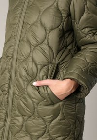 Olijfgroene gewatteerde pufferjack met een ritssluiting aan de voorkant, groot circulair stikpatroon, zijzakken en een zachte voering bij de manchetten.