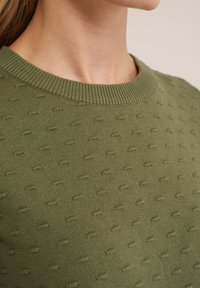 Close-up of a person wearing an olive green knitted sweater with a textured dot pattern and ribbed crew neckline.