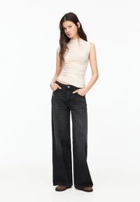 Top senza maniche beige con colletto alto, abbinato a jeans neri a gamba larga con chiusura a bottone e tasche laterali.