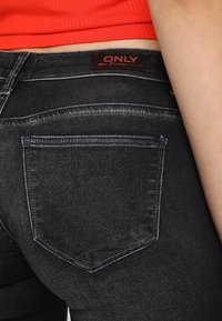 Los vaqueros de mezclilla negra presentan un bolsillo trasero con un sutil efecto de desgaste. La cinturilla muestra la etiqueta de la marca "ONLY" en un color contrastante.