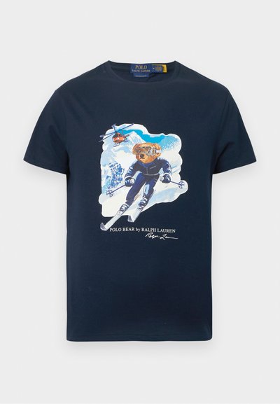 Tamnoplava majica od pamuka s grafikonom medvjeda na skijanju u skijaškoj opremi, s helikopterom u pozadini i imenom marke ispod.