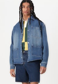 Dark blue denim jacket with large front pockets, button closure, and relaxed fit, layered over a striped green and yellow polo.
