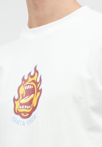 Un tricou alb din bumbac prezintă un grafic mare cu o gură în flăcări, cu culorile roșu, galben și albastru, și textul "SANTA CRUZ" dedesubt.