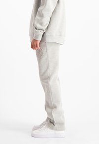 Persoon in lichtgrijze sweatpants en bijpassende sweatshirt, met witte Nike Air Force 1 sneakers, staande in profiel op een witte achtergrond.