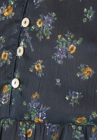 Tissu noir avec de petits motifs floraux en jaune, violet et vert. Comprend des boutons couleur crème et un ourlet froncé.