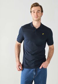 Navy plava kratka polo majica s tri gumba i malim žutim logotipom na prsima, uparena s svijetloplavim trapericama.
