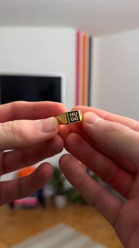 Guldring med flat yta; ingraverad text "HU GO" i svart på ena sidan. Slät textur, enkel design, hålls mellan fingrarna.