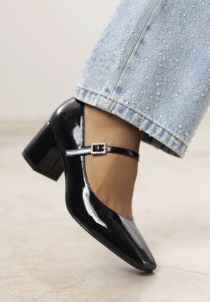 Scarpe Mary Jane in vernice nera con tacco a blocco, caratterizzate da una larga fascia e un dettaglio con fibbia. Posizionate accanto a un denim azzurro sbiadito.