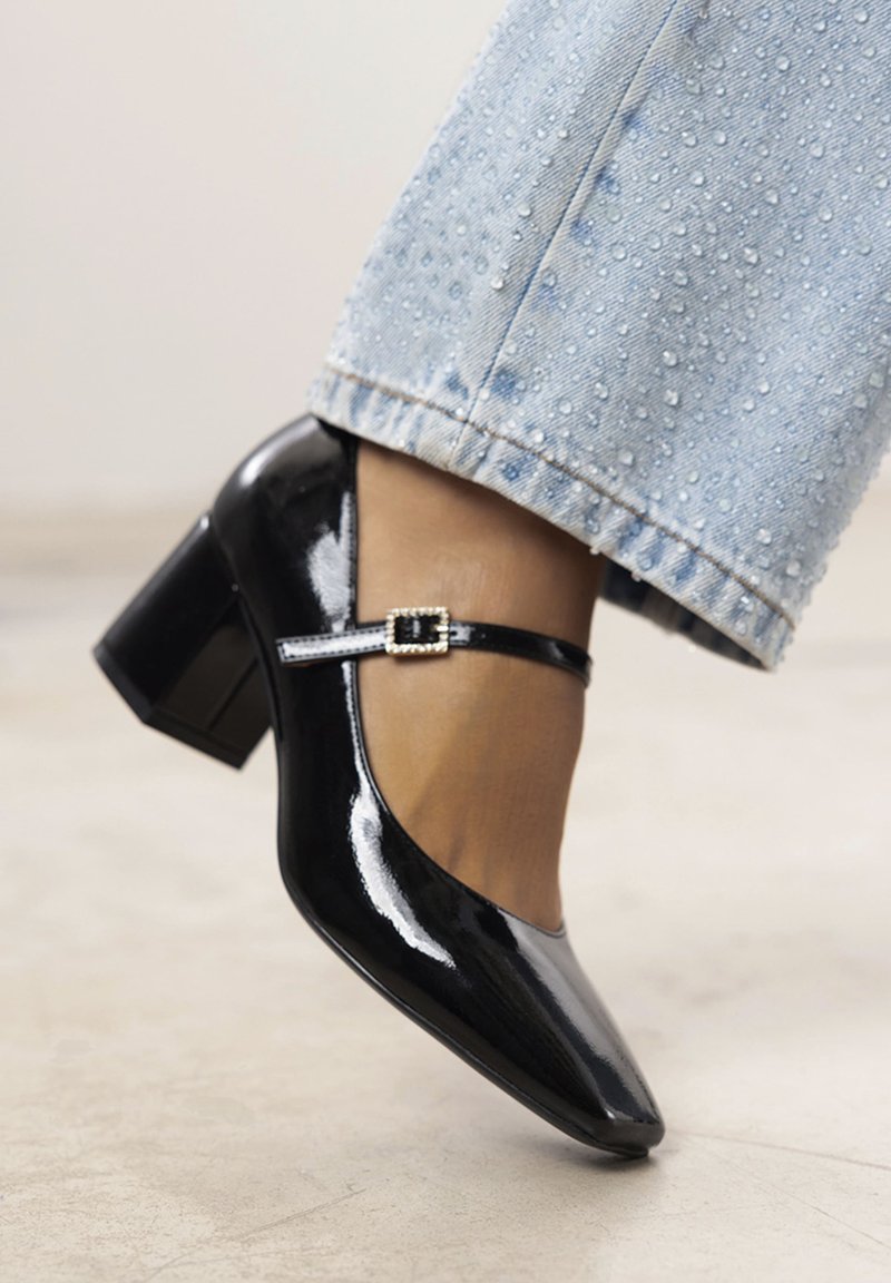 Scarpe Mary Jane in vernice nera con tacco a blocco, caratterizzate da una larga fascia e un dettaglio con fibbia. Posizionate accanto a un denim azzurro sbiadito.