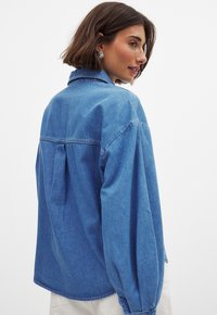 Denim jacket in light blue, featuring a relaxed fit, long sleeves, and a back pleat detail. Smooth texture and classic collar design.