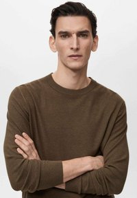 Brauner, strukturierter Strickpullover mit Rundhalsausschnitt und langen Ärmeln, getragen von einer Person mit verschränkten Armen, vor einem neutralen Hintergrund.