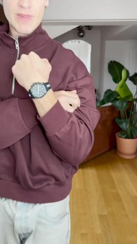 Black watch with silver accents on a wrist, worn over a maroon zip-up jacket. Light-colored jeans visible, with green plants in the background.