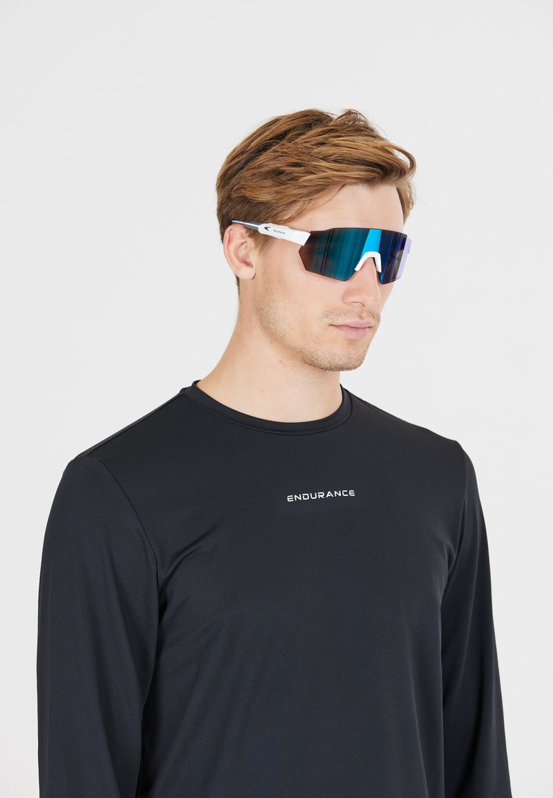 Man in een zwart langslievend sportshirt met de tekst "ENDURANCE" en reflecterende sportzonnebril met witte frames, die naar rechts kijkt.