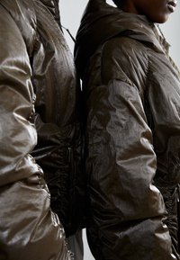 Puffjackor i mörkbrunt, med en glänsande, texturerad yta, huva och elastiska fållar. Synlig dragkedjeförsedd detalj på sidan.