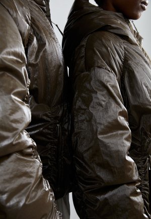 Pufferjassen in donkerbruin, met een glanzend, textuur oppervlak, capuchon en elastische boorden. Zichtbare ritsdetails aan de zijkant.
