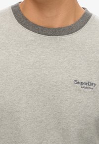 Lichtgrijze katoenen sweatshirt met een ribbelboord aan de grijze ronde hals en een geborduurd "SuperDry Athletics" logo in donkerblauw op de borst.