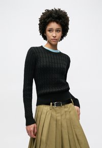 Pull noir en maille avec texture torsadée, col rond et poignets côtelés porté sur une chemise bleu clair. Associé à une jupe plissée beige.