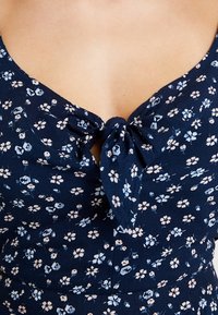 Námořnické květinové šaty s malými bílými a světle modrými květy, s propleteným předním detailem a přiléhavým korzetem. Měkká textilie.