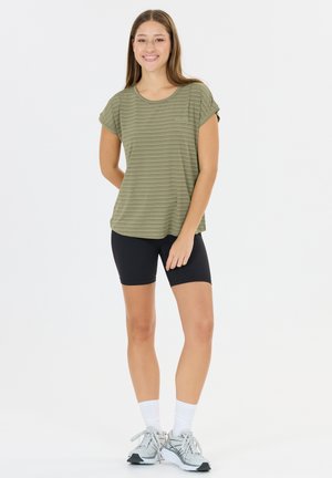 Nuori nainen seisoo, hänellä on päällään vihreäraidallinen lyhythihainen paita, mustat pyöräilyshortsit, valkoiset sukat ja harmaat juoksukengät, hymyilee eteenpäin.