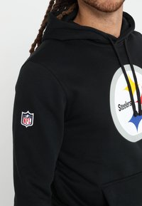 Sudadera con capucha negra con un logo circular que incluye la palabra "Steelers" y un emblema a color. Incorpora el logo de la NFL en la manga. Tejido suave y diseño casual.
