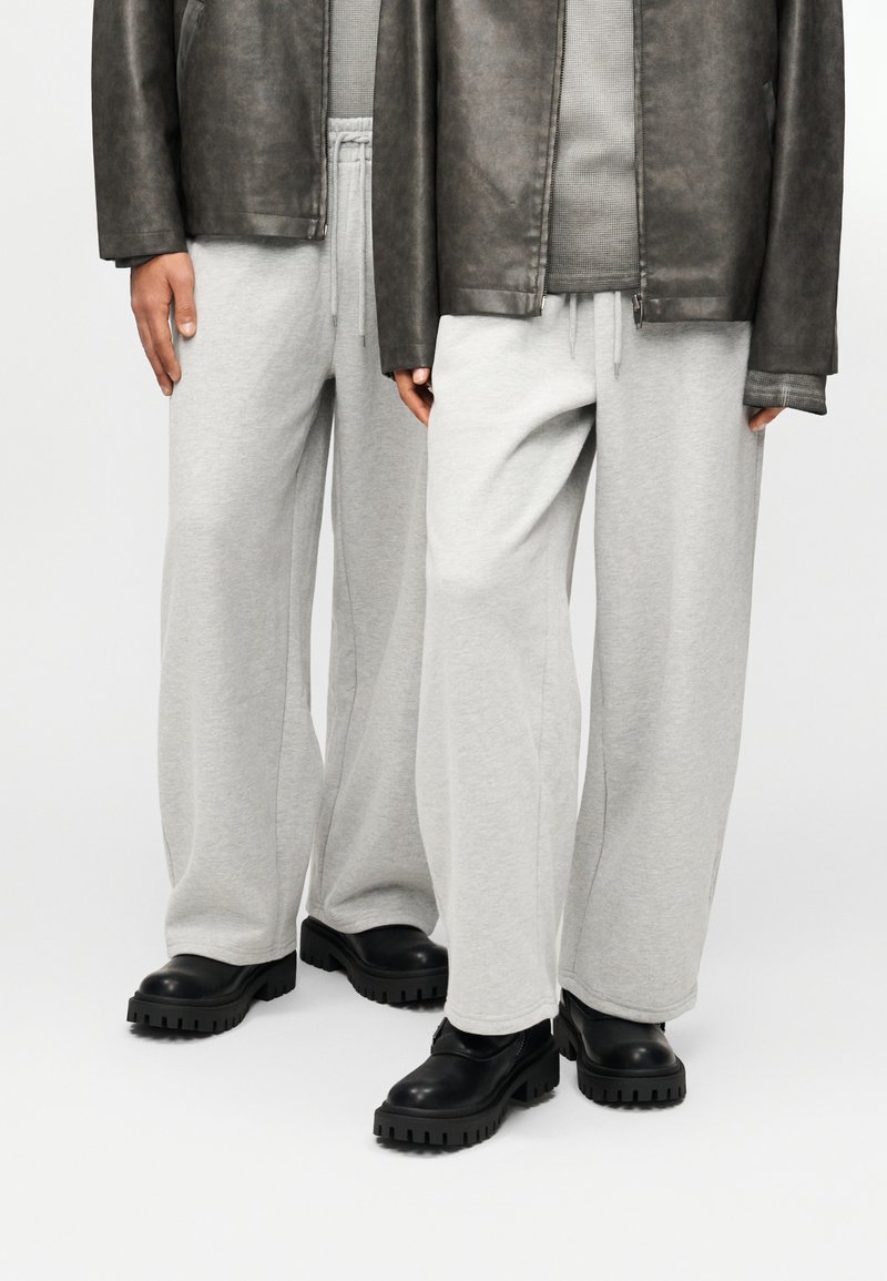 Γκρι φόρμα με άνετη εφαρμογή και ελαστική μέση, σε συνδυασμό με μαύρα παπούτσια με χοντρή σόλα. Το φόντο είναι απλό λευκό.